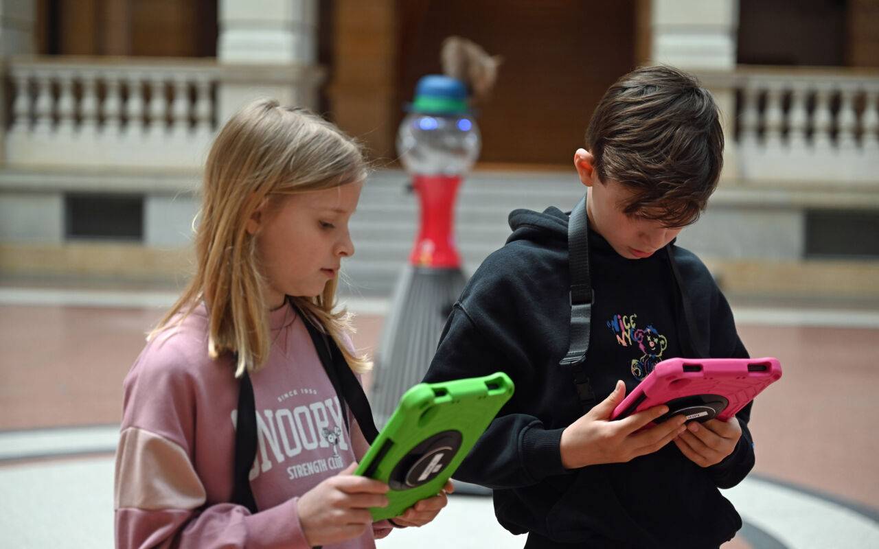 Linus (r) und Matilda stehen mit Tablets vor einem Roboter im Museum für Kommunikation Berlin.