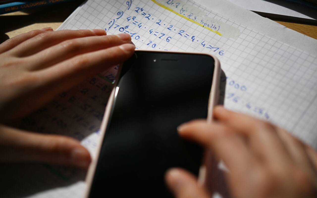 Ein Kind hält ein Smartphone über einem Blatt mit Mathematikaufgaben - das Gerät liegt auf dem Arbeitsblatt.