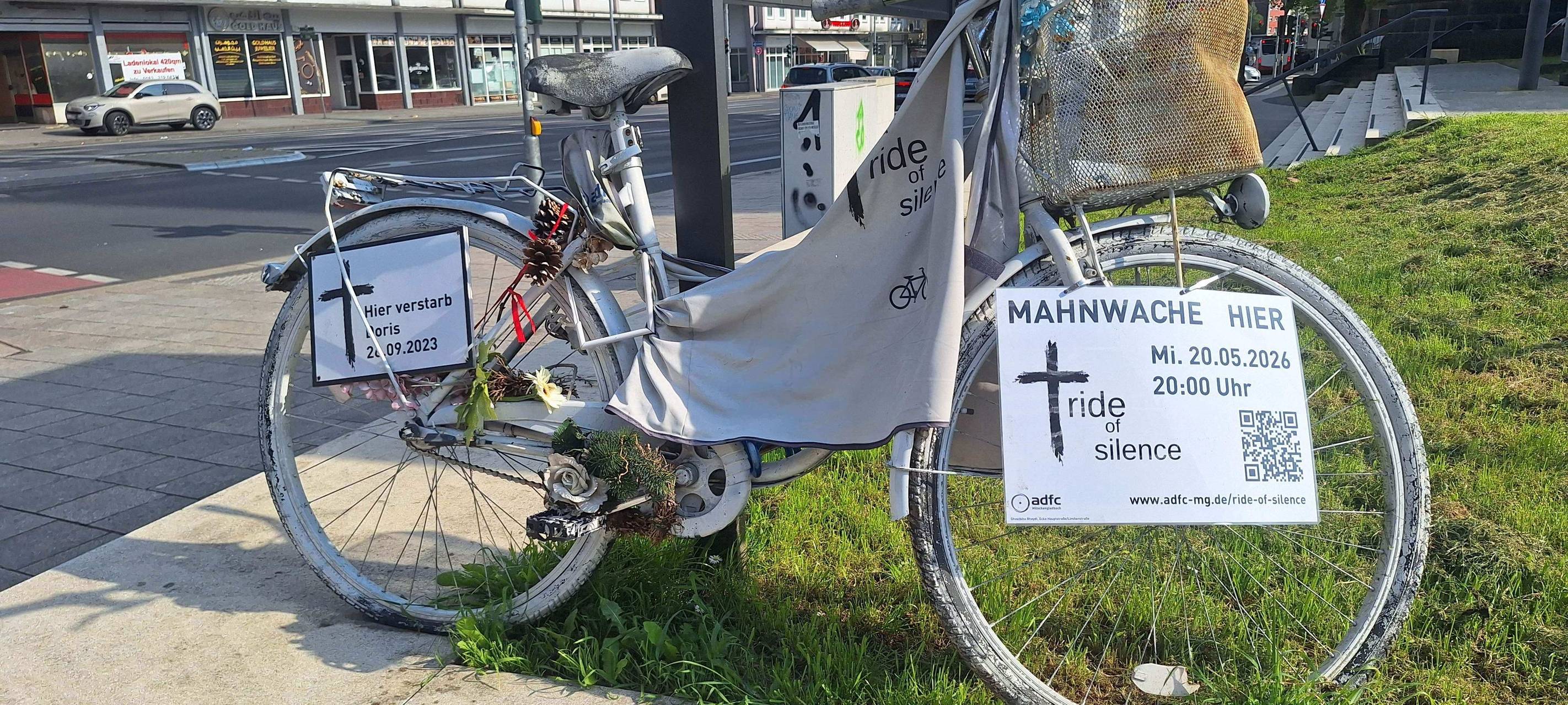 Ein weißes Fahrrad mit Schildern. Auf den Schildern ist ein Nachruf zu sehen. Außerdem eine Ankündigung zu einer Mahnwache.